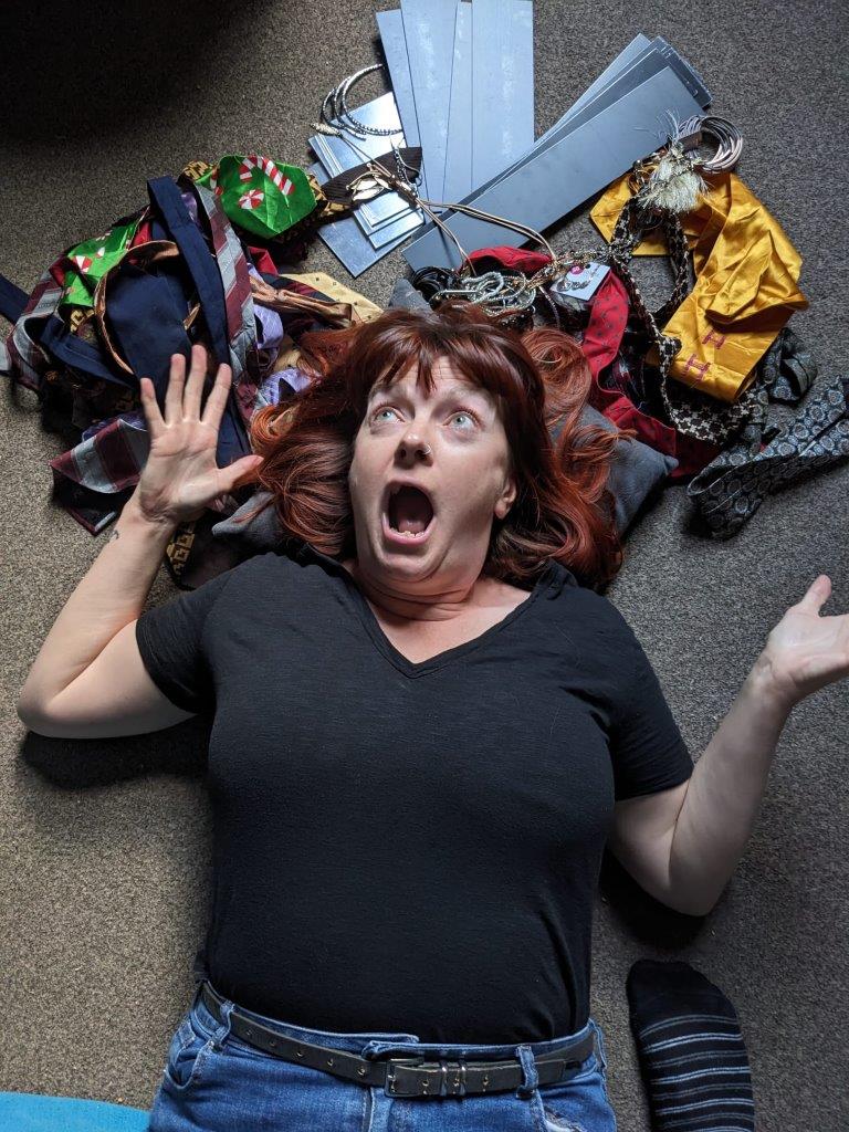 A white woman with dark hair is lying on the ground wearing a black tshirt and blue jeans. She is surrounded by waste textiles that she'll make into jewellery and has a shocked look on her face.