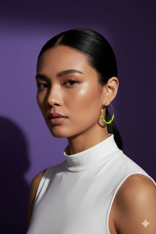 A woman wearing pair of silver tone hoop earrings with yellow and green beads made from upcycled materials, on a purple background