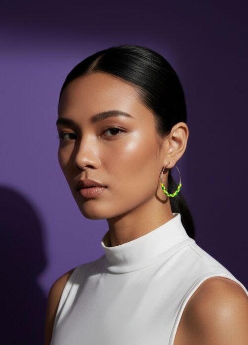 A woman wearing pair of silver tone hoop earrings with yellow and green beads made from upcycled materials, on a purple background