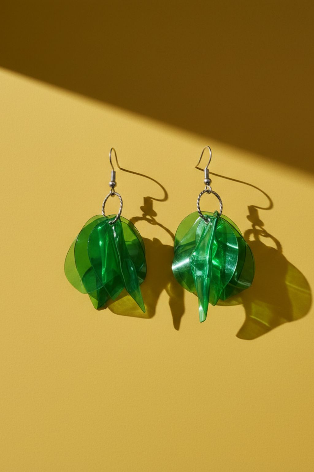 A pair of silver tone earrings with green charms, upcycled from waste plastic on a mustard yellow background