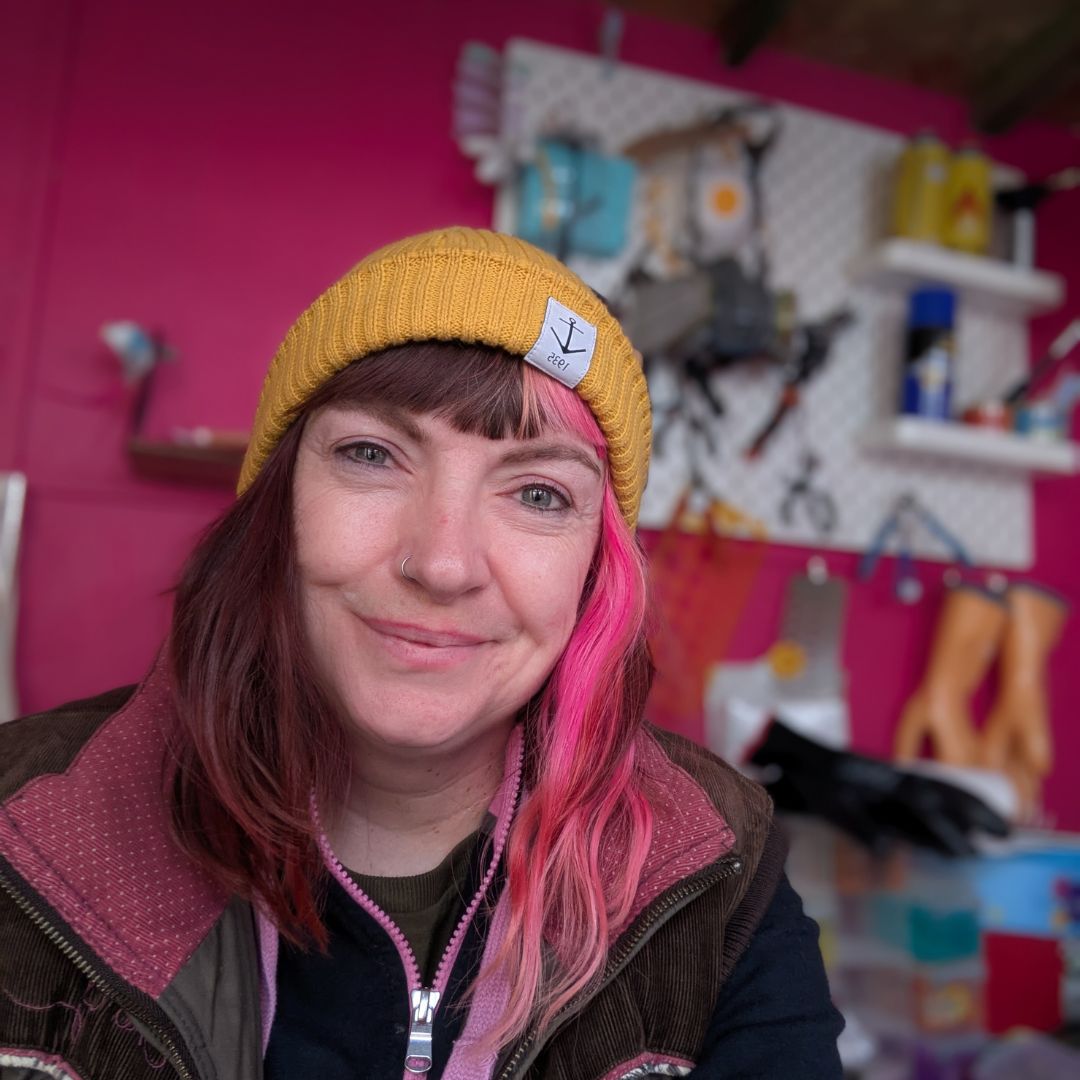 A white woman with pink hair and a yellow hat poses for the camera in her workshop. The walls are pink and tools are hanging up. Trash Panda founder creating sustainable jewellery from reclaimed materials in the UK