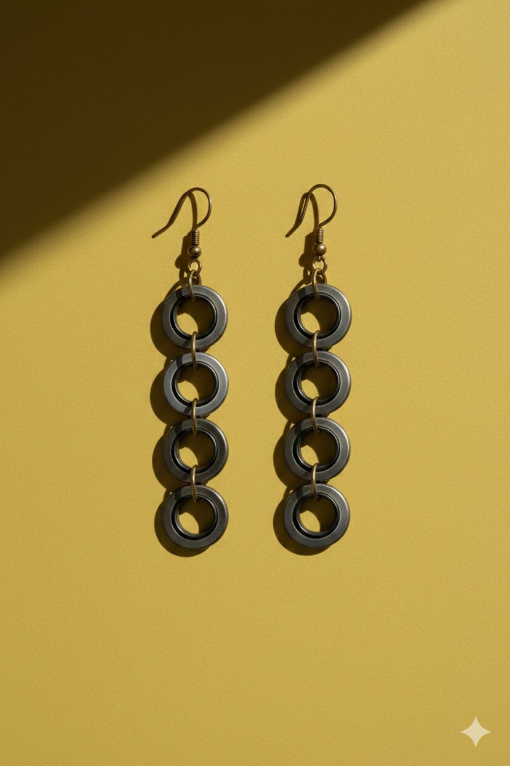 A pair of industrial-style earrings made from upcycled materials, they are dangly with 4 small rings hanging. On a mustard yellow background