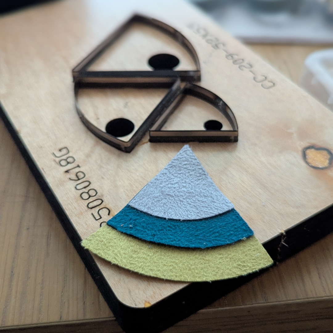 Three layered triangles of material, in different colours, sit on top of a leather cutting press ready to be made into jewellery