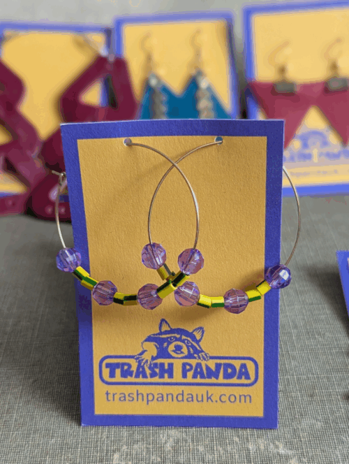 Upcycled hoop earrings with yellow-and-green electrical wire beads and faceted purple beads on stainless steel hoops, displayed on Trash Panda card