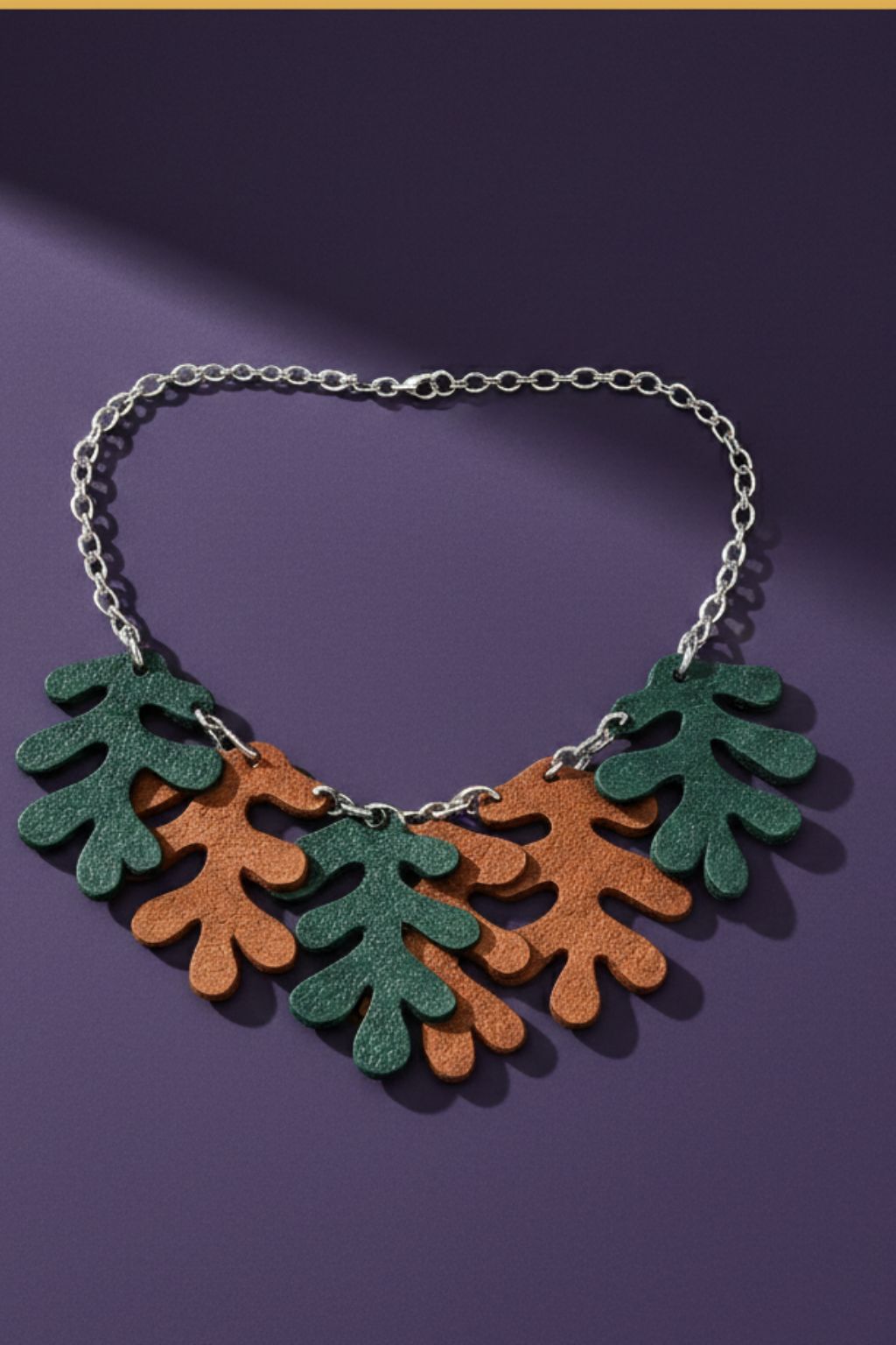 A necklace made from a silver tone chain and oak-shaped leaves cut from leather, on a purple backgound