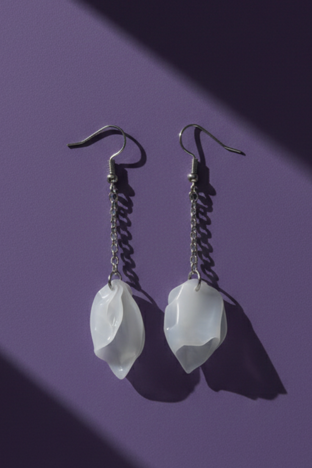 A pair of silver tone earrings with a dangling chain and white petals at the bottom. Made from upcycled plastic, on a purple background.