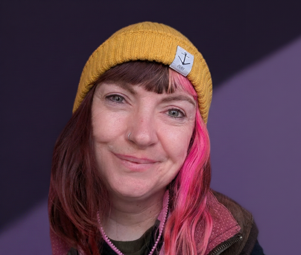 A white woman with a pink streak in her hair looks at the camera. She is wearing a mustard coloured beanie hat and is against a purple background